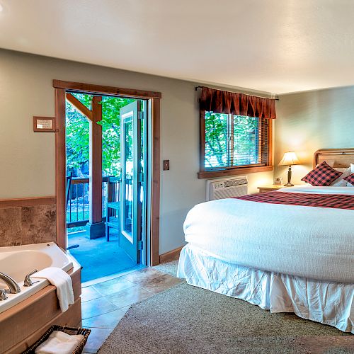 A cozy bedroom with a large bed, nightstands, lamps, a framed picture, a jetted tub, and a doorway leading to a porch with greenery outside.