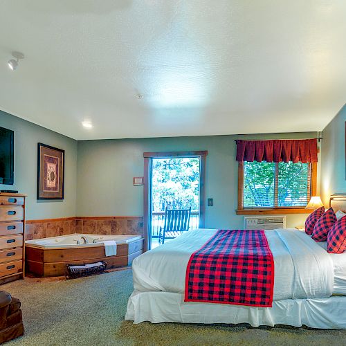 A cozy bedroom with a large bed, a plaid blanket, a flat-screen TV, a dresser, and a wooden chair. French doors lead to the outside.