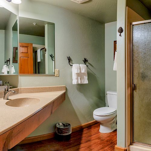 A bathroom features a large sink, mirror, toilet, towel hooks, trash can, and a glass-enclosed shower with a towel over the top.