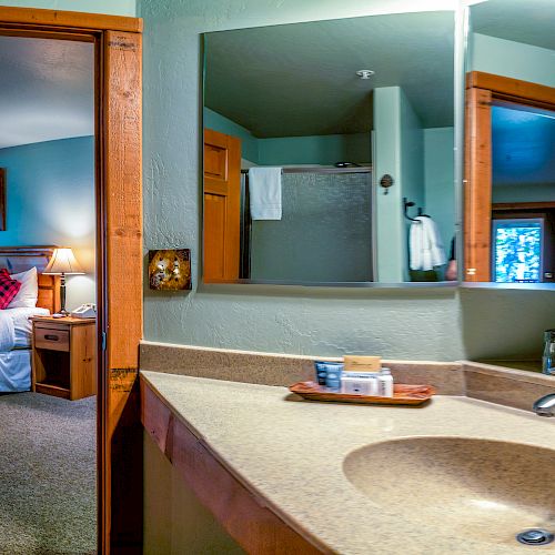 The image shows a cozy bedroom with a bed, wooden furniture, and a view of a bathroom sink and mirror, reflecting the shower and towels.