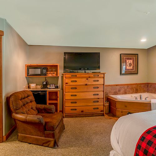 A cozy bedroom with a bed, chair, TV, microwave, dresser, a jacuzzi, and an open bathroom door leading to a shower and toilet.