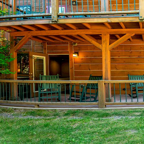 A wooden cabin with a porch, several chairs, and greenery in the foreground. The structure has two levels with a balcony on top.