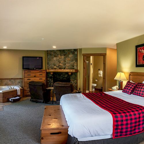 A cozy hotel room features a large bed with a red and black checked runner, a fireplace, TV, desk, armchair, hot tub, and framed artwork.
