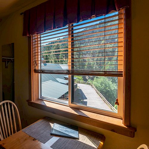 A small table and chair are placed by a window with blinds, overlooking rooftops and trees; a book lies on the table, catching sunlight.