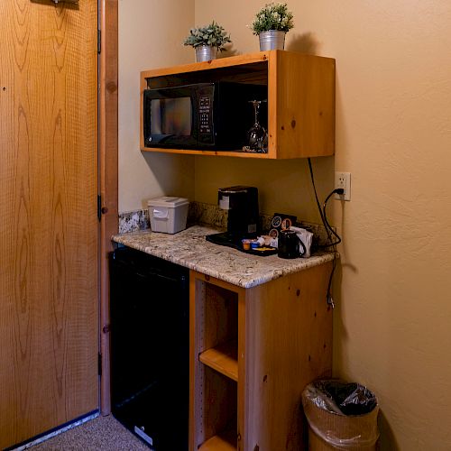 A kitchenette with a microwave, coffee maker, mini-fridge, plants on a shelf, and a trash can is beside a wooden door, with some appliances and cords.