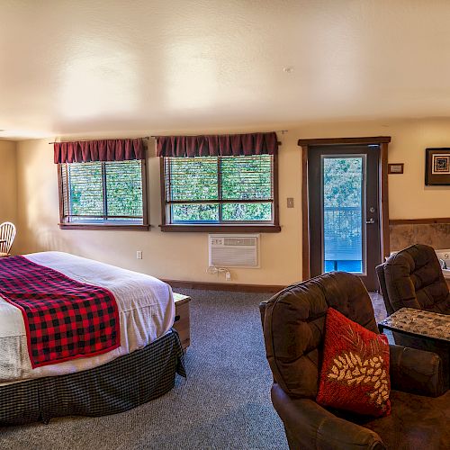 This image shows a cozy bedroom with a large bed, armchairs, a dresser, TV, and a doorway to a balcony. The room has a warm, inviting ambiance.