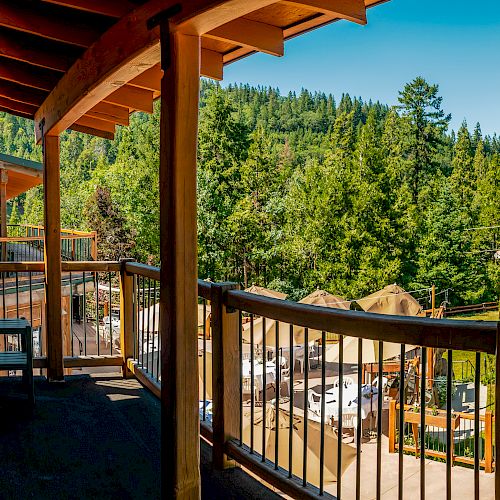 A wooden deck overlooking a green forested area, with several chairs and tables along the railing. Mountains and trees in the background.