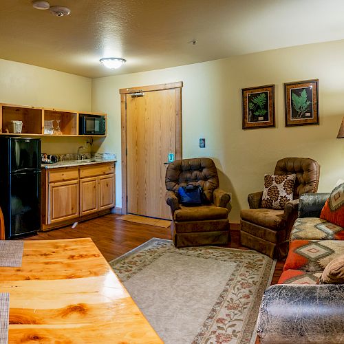 A cozy living area with a sofa, two armchairs, a small kitchen, a wooden dining table, and a TV. The room is warmly lit and decorated.