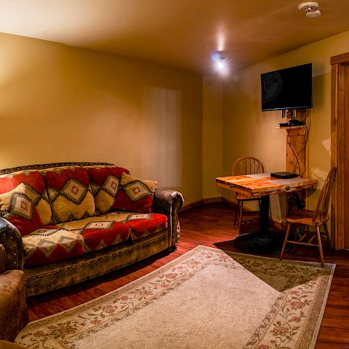 A cozy living room features patterned furniture, a wall-mounted TV, a small wooden table with chairs, a lamp, and an area rug, all warmly lit.