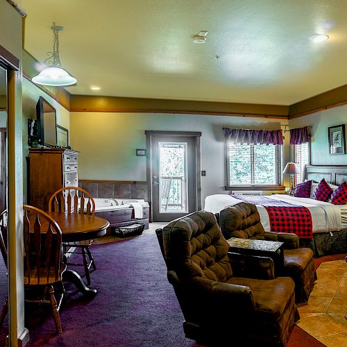 A cozy hotel room features two beds, a seating area with armchairs, a dining table, a mirrored wardrobe, and a window with a scenic view.