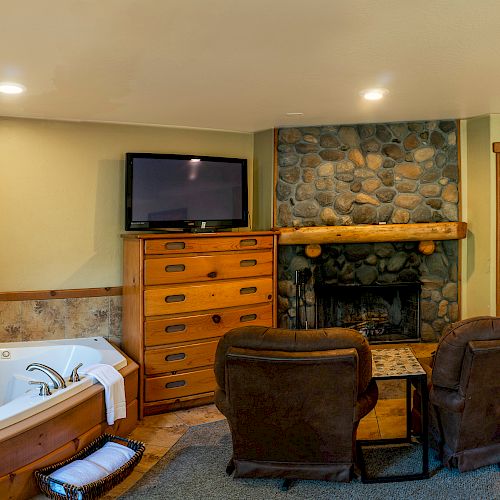 A cozy room with a hot tub, TV, stone fireplace, two chairs, and a small table, leading to a bathroom and entryway.