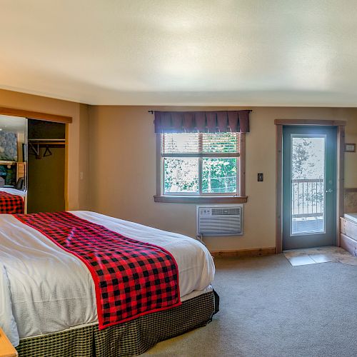 A cozy bedroom with a large bed, checkered bedding, a lamp on the nightstand, an armchair, a jacuzzi, and a door with a view of trees is shown.