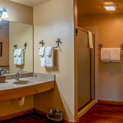 A bathroom with a double sink, mirror, shower, toilet, towel racks with white towels, wooden flooring, and a wicker basket is shown.