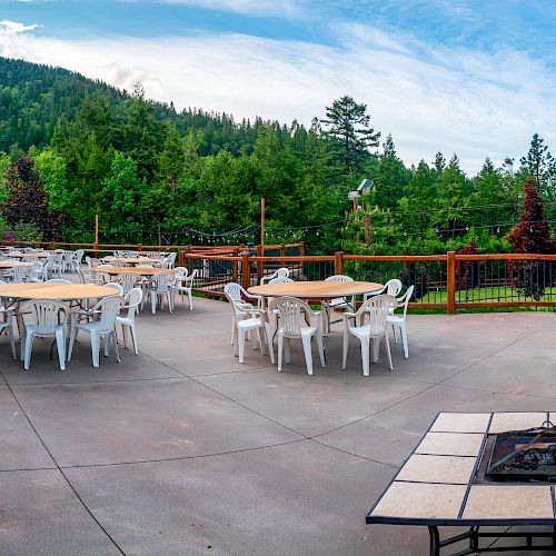 This image shows an outdoor patio area with round tables and white chairs, surrounded by a wooden fence and forest scenery.