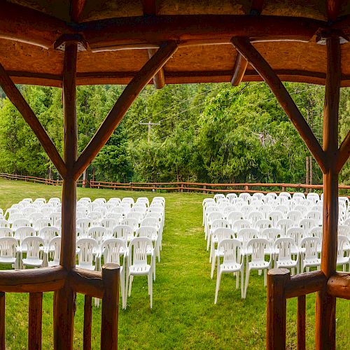 Several rows of white chairs are arranged on a grassy area, viewed from under a wooden gazebo, with trees and lush greenery in the background.