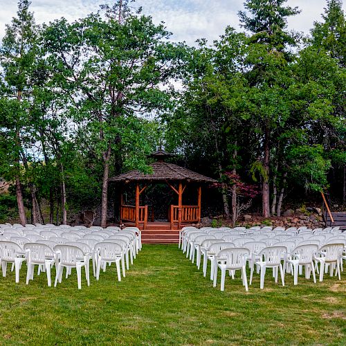 An outdoor ceremony setup with white chairs facing a wooden gazebo, surrounded by trees and greenery, likely for a wedding or event.