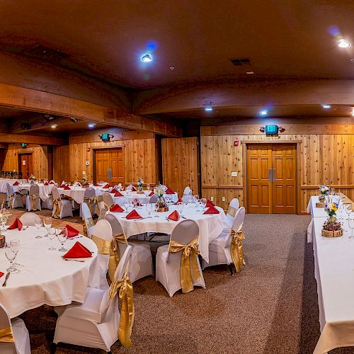 The image shows a banquet hall with round tables set for dining, each with white tablecloths, red napkins, and golden chair sashes.