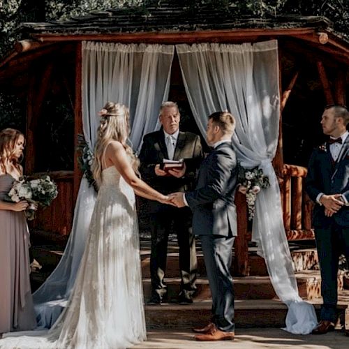 A couple is exchanging vows in an outdoor wedding ceremony, with the officiant and two attendants standing nearby on a wooden platform with drapes.