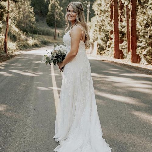 A bride in a white wedding dress stands on a road holding a bouquet, with tall trees lining the background and sunlight filtering through.