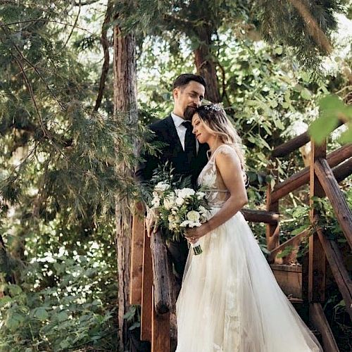 A bride and groom stand close together on wooden stairs amidst greenery. The bride holds a bouquet, and the couple looks happy and serene in the moment.