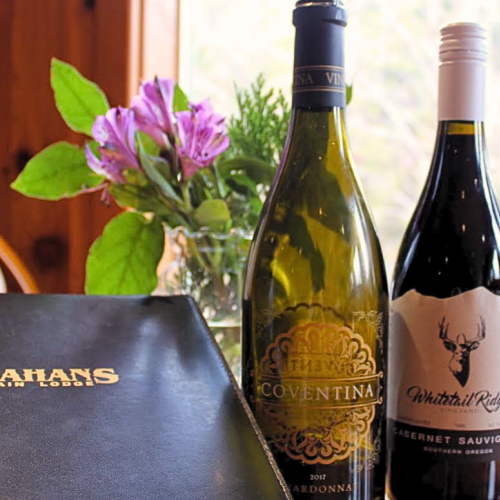 The image shows a leather-bound menu, three bottles of wine, and a vase with purple flowers on a wooden table near a window.