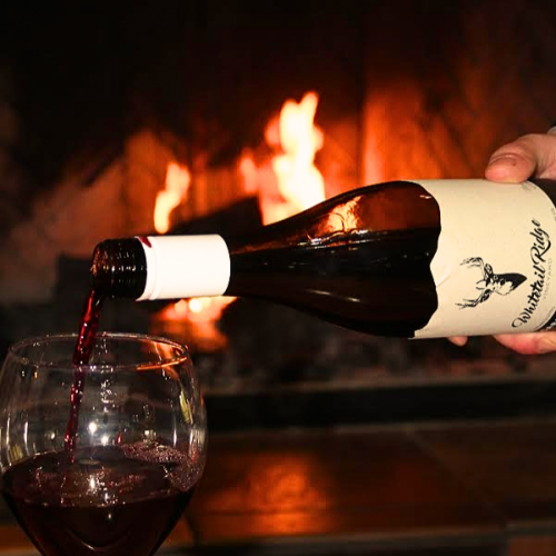 A hand pours red wine from a bottle into a glass in front of a fireplace, with flames burning in the background.