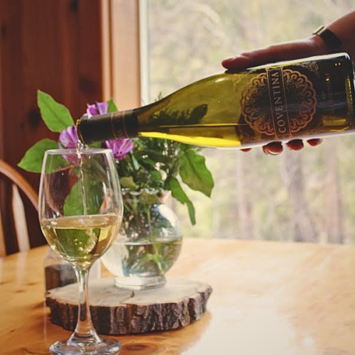 A hand is pouring white wine from a bottle into a wine glass on a wooden table. A vase with flowers is placed in the background.