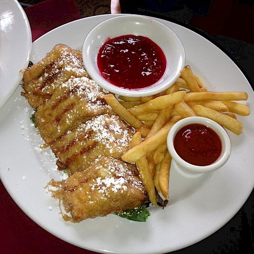 The image shows a plate of fried food, including deep-fried items dusted with powdered sugar, French fries, and two dipping sauces in small bowls.