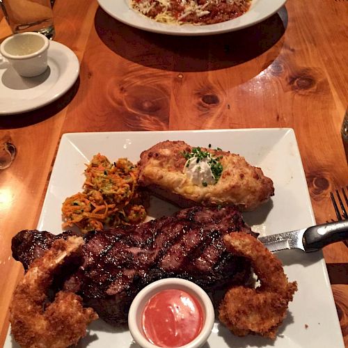 A wooden table with a steak, shrimp, roasted potato, and dipping sauce on a square plate. In the background, there's a pasta dish on a round plate.