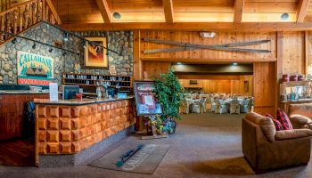A rustic wooden interior features a reception desk, seating area, signage, and a cozy dining space with tables set up in the background.