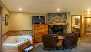 The image shows a cozy room with a fireplace, a TV on a wooden dresser, two armchairs, and a jacuzzi. A bathroom is visible in the background.