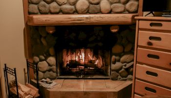 A cozy stone fireplace with a wreath above, flanked by chairs and a wooden dresser, creates a warm and inviting atmosphere.