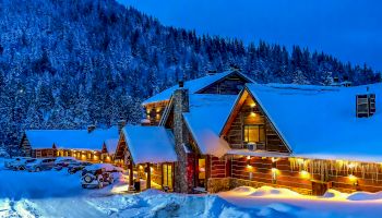 A cozy, snow-covered lodge with warm lighting sits in a mountainous forest area during twilight. Cars are parked outside, and snow blankets the scene.