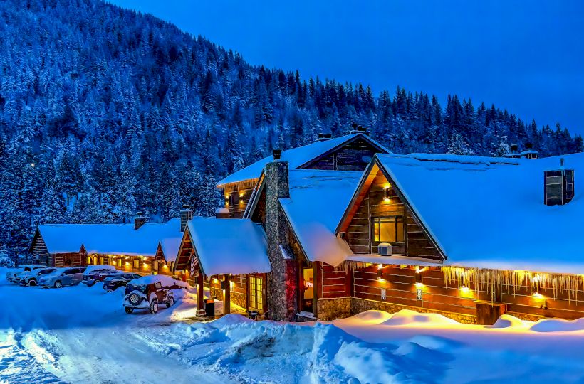 A cozy, snow-covered lodge with warm lighting sits in a mountainous forest area during twilight. Cars are parked outside, and snow blankets the scene.