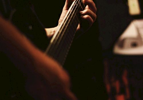 A person is playing an electric guitar in a dimly lit setting, focusing on their hand gripping the neck of the guitar and pressing the strings.