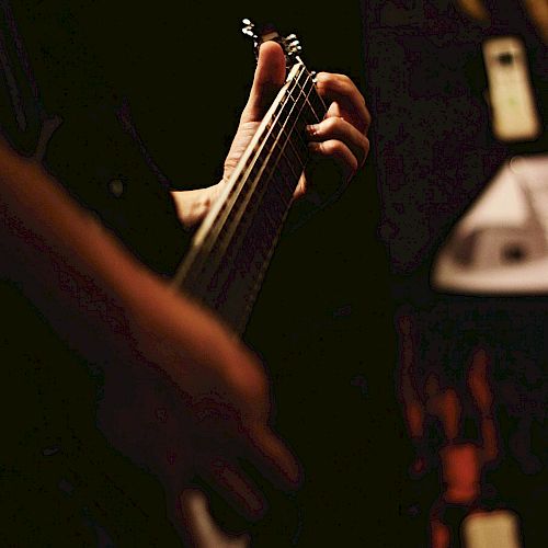 A person is playing an electric guitar in a dimly lit setting, focusing on their hand gripping the neck of the guitar and pressing the strings.