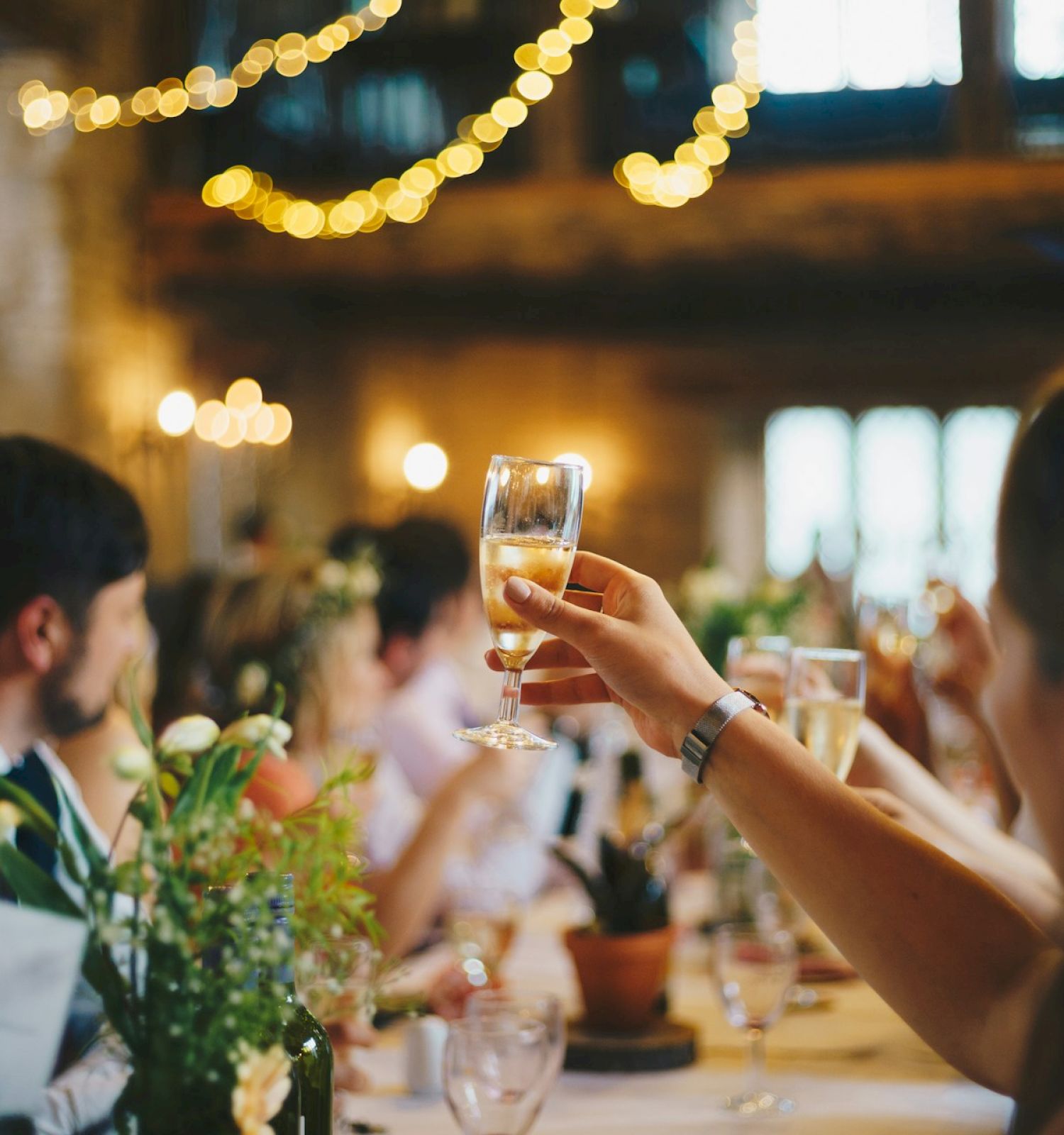 People are raising glasses in a celebratory toast at an indoor event, with flowers and decorations visible on the tables.