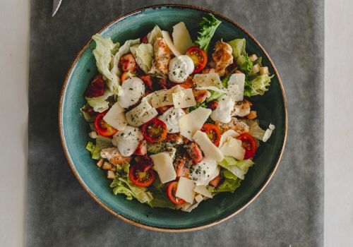 The image shows a colorful salad with cherry tomatoes, lettuce, cheese, and herbs in a blue bowl on a gray placemat.