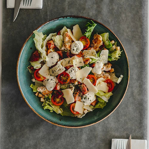 The image shows a colorful salad with cherry tomatoes, lettuce, cheese, and herbs in a blue bowl on a gray placemat.