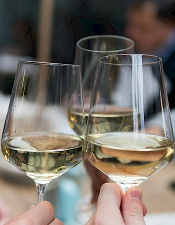 Two people clinking glasses of white wine at a table in a blurred restaurant setting, celebrating or toasting together.