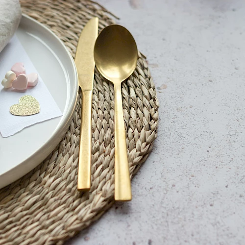 A stylish place setting with a white plate, gold cutlery, a woven placemat, and small decorative cards with hearts, plus flowers.