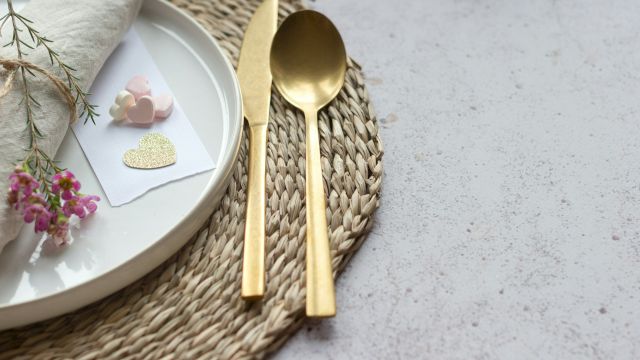 A stylish place setting with a white plate, gold cutlery, a woven placemat, and small decorative cards with hearts, plus flowers.