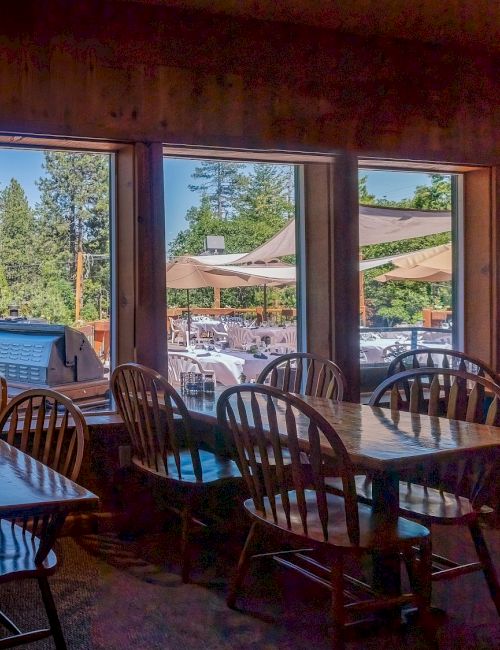 A cozy dining room with wooden tables and chairs, large windows showing outdoor trees and umbrellas, bathed in warm natural light.