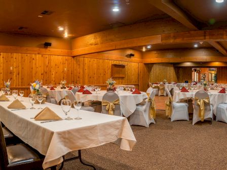 A cozy banquet hall with wooden walls, round tables draped in white linens, gold chair sashes, and neatly set place settings for a formal event.