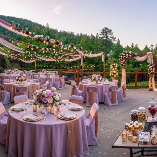 An outdoor wedding reception with round purple-draped tables, floral centerpieces, string lights, and a dessert bar under a wooden decked venue.