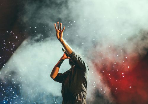 A performer on stage raises one hand amid colorful smoke and confetti, creating a dramatic live-show vibe.