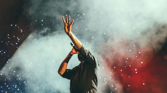 A performer on stage raises one hand amid colorful smoke and confetti, creating a dramatic live-show vibe.