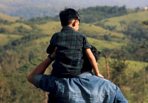 A man carries a child on his shoulders, overlooking rolling hills and distant mountains in a lush, panoramic landscape.