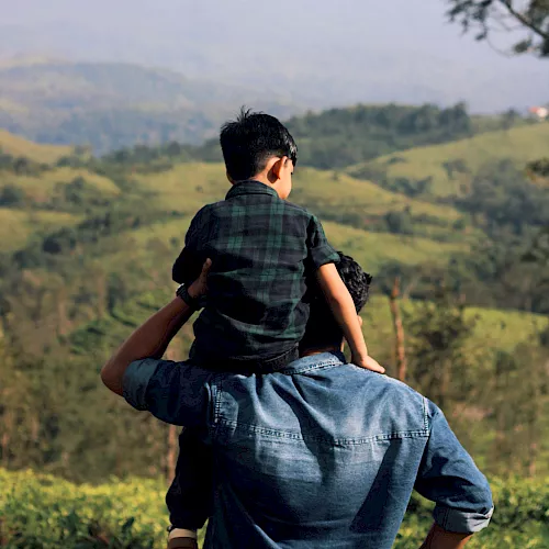 A man carries a child on his shoulders, overlooking rolling hills and distant mountains in a lush, panoramic landscape.