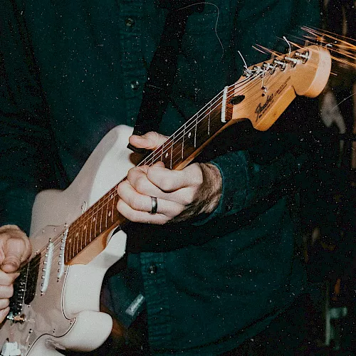 A guitarist&rsquo;s hands playing an electric guitar, fingers on the strings while bright light streaks cross the scene.
